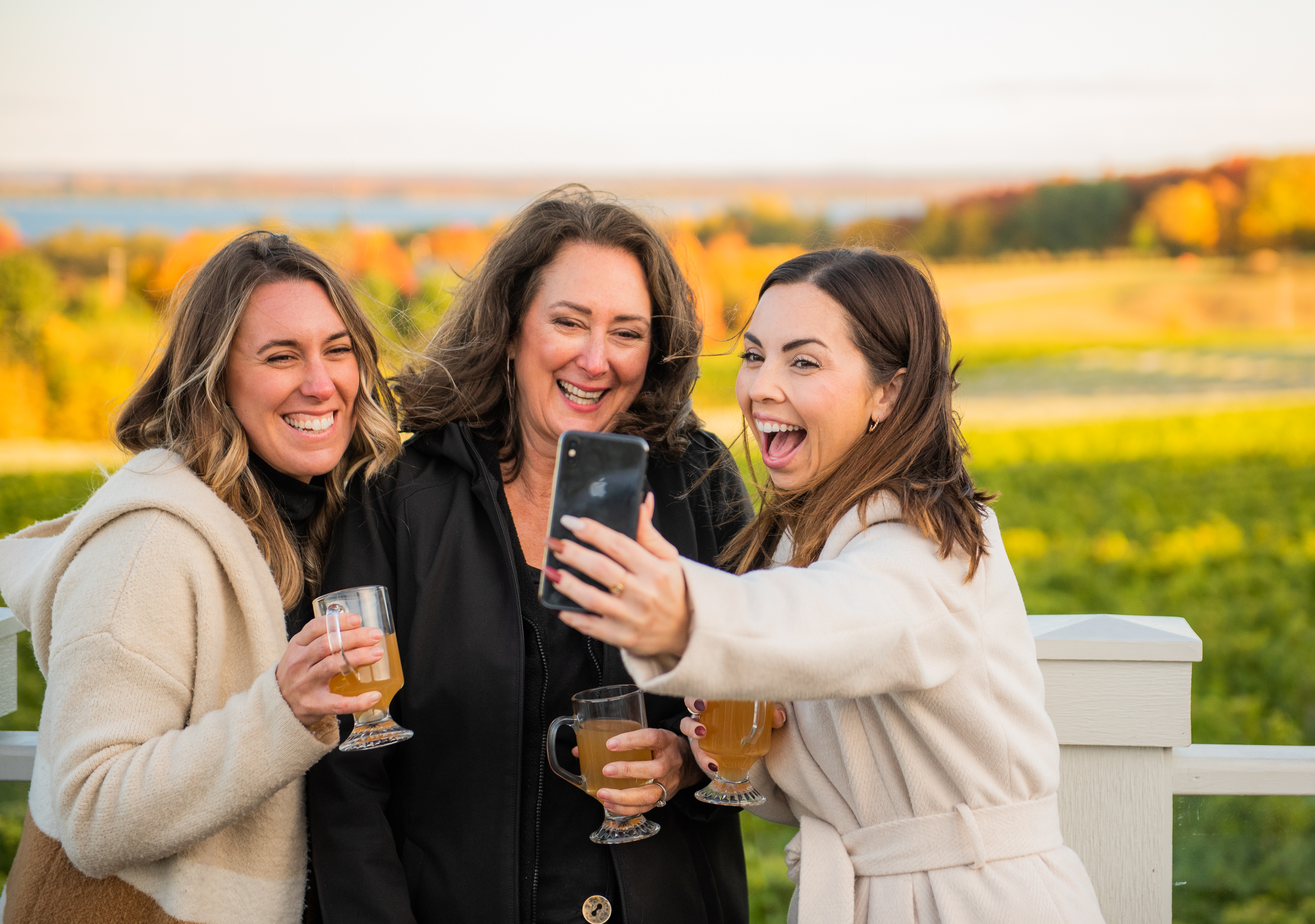 Friends taking selfie outside with drinks in hand and color trees in the distance behind them